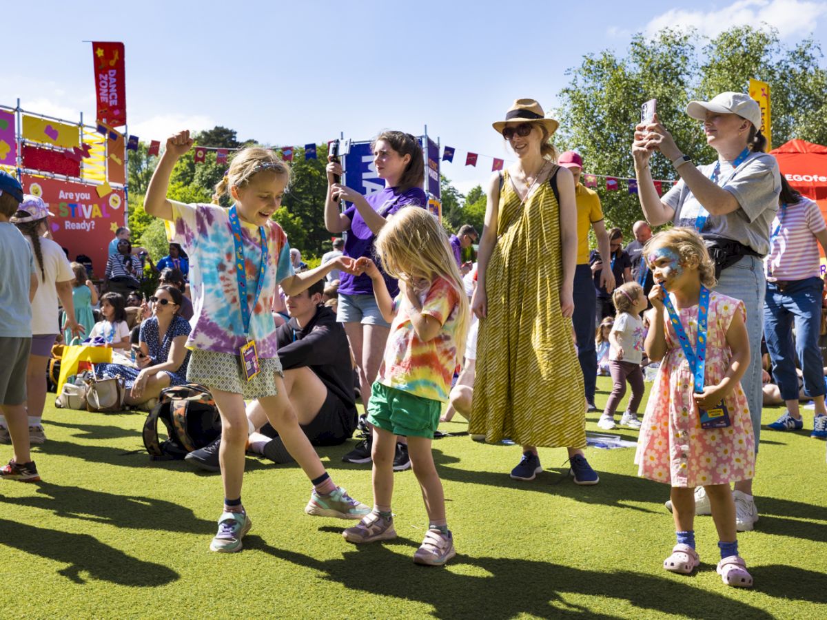 Kids with face paint dance at an outdoor festival on a sunny lawn, while parents and flags and tents fill the background.