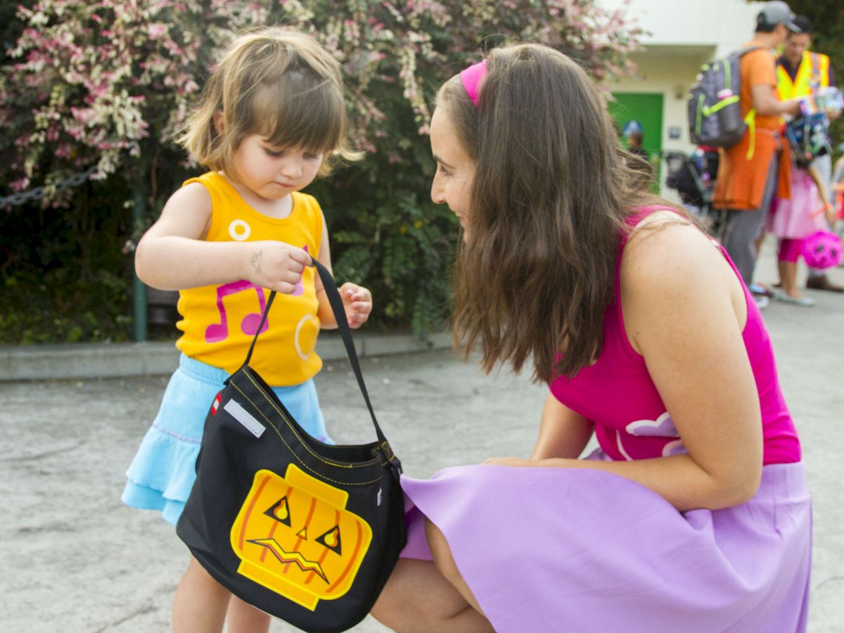 A caregiver chats with a little child outdoors as the kid clutches a Halloween-style bag near colorful flowers and park goers.