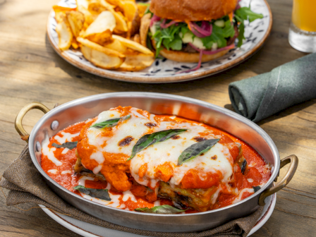 A cheesy chicken parm skillet with melted mozzarella and tomato sauce, plus a burger, fries, and a glass of orange drink on the table.