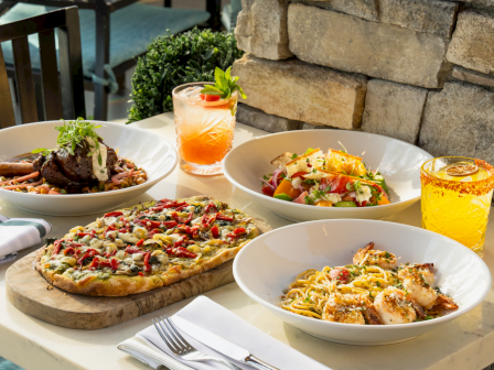 An outdoor table spread with pizza, pasta, salad, and meat dishes, plus citrus drinks in cut-glass tumblers and a lemon garnish!