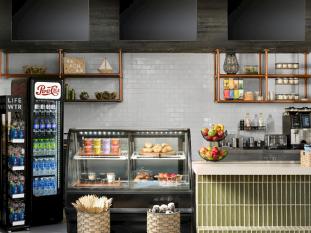 A cozy caf&eacute; counter setup with a glass bakery display, a glowing drink fridge, shelves of snacks, and a fruit bowl.