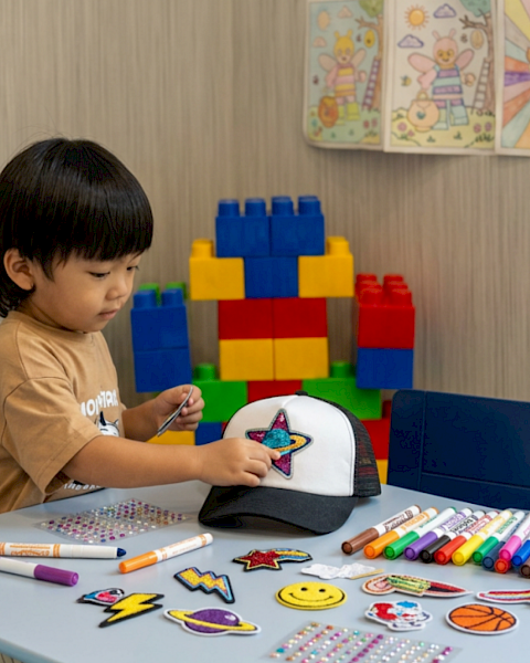 A toddler decorates a star patch on a cap with markers at a table, with colorful building blocks in the background.