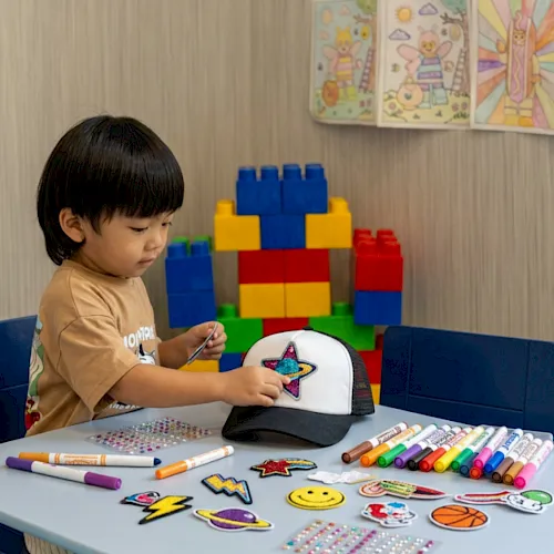 A toddler decorates a star patch on a cap with markers at a table, with colorful building blocks in the background.