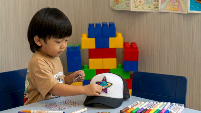 A toddler decorates a star patch on a cap with markers at a table, with colorful building blocks in the background.