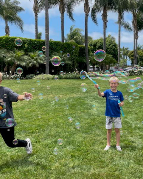 Kids play in a sunny park blowing soap bubbles, with tall palm trees and lush hedges in the background.