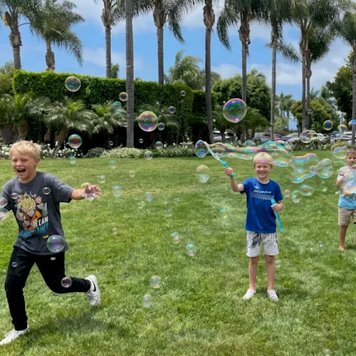 Kids play in a sunny park blowing soap bubbles, with tall palm trees and lush hedges in the background.