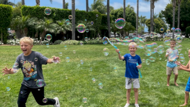 Kids play in a sunny park blowing soap bubbles, with tall palm trees and lush hedges in the background.