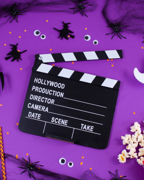 A purple Halloween film set with a clapperboard, spiders and ghost stickers, popcorn, striped candy, and film snacks all around.