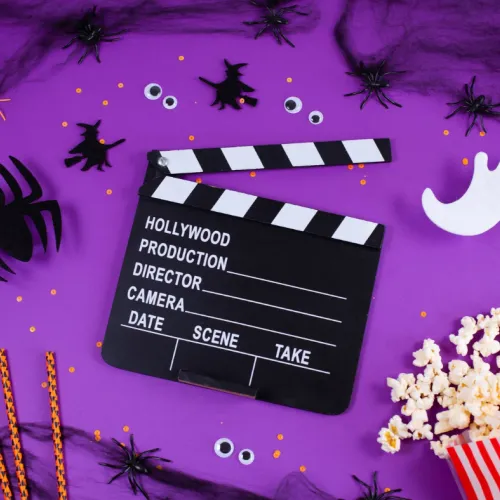 A purple Halloween film set with a clapperboard, spiders and ghost stickers, popcorn, striped candy, and film snacks all around.