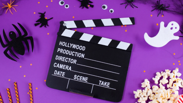 A purple Halloween film set with a clapperboard, spiders and ghost stickers, popcorn, striped candy, and film snacks all around.