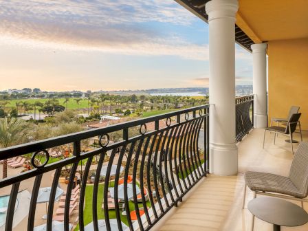 Balcony with cushioned chairs and railing, overlooking landscaped gardens, palm trees, a pool, and distant ocean at sunset.