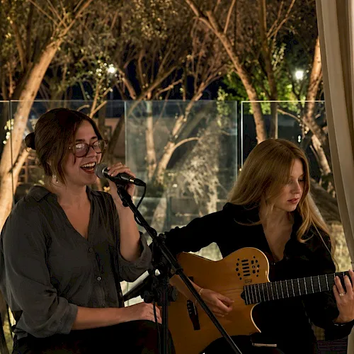 Two women perform live music outdoors at dusk, one singing into a microphone while the other plays an acoustic guitar.