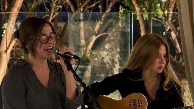 Two women perform live music outdoors at dusk, one singing into a microphone while the other plays an acoustic guitar.