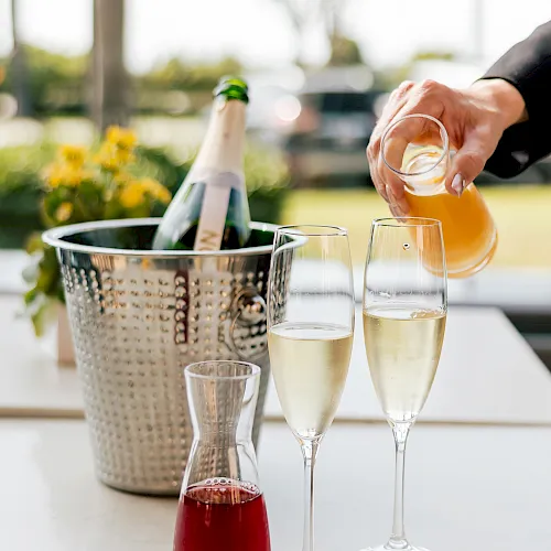A person pours sparkling champagne into flutes on an outdoor patio, with a pitcher of orange drink and flower decor in view.