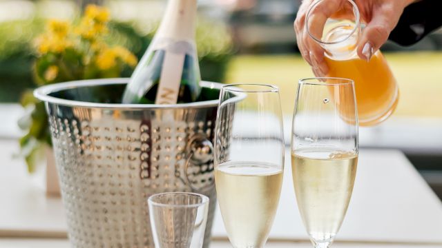 A person pours sparkling champagne into flutes on an outdoor patio, with a pitcher of orange drink and flower decor in view.